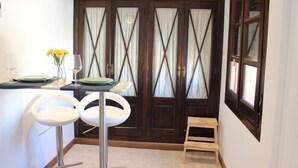 Un'area pranzo con un tavolo apparecchiato per due, due sgabelli bianchi, uno sgabello di legno e un vaso con fiori gialli.