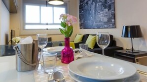 Un coin repas avec une table dressée pour deux personnes, un vase rose orné de fleurs et un tableau représentant un paysage urbain accroché au mur.