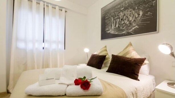Un lit bien fait, avec du linge de lit blanc, une rose rouge et une serviette accompagnée d’une carte.