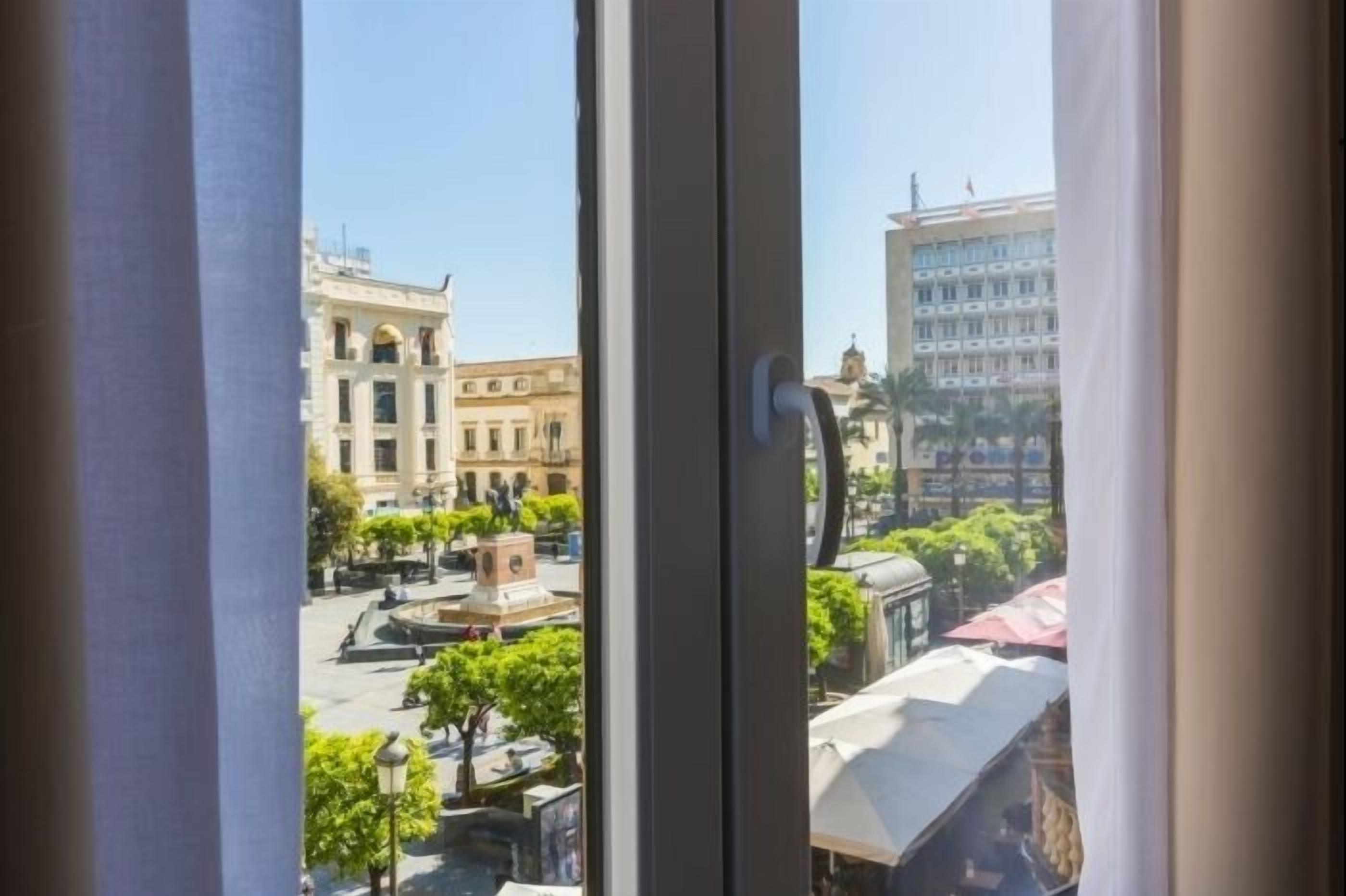A view from a window with a view of a plaza, buildings, and a fountain.