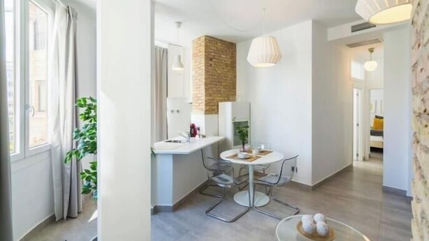 Habitació moderna i ben il·luminada amb zona de cuina, taula de dinar i un gran finestral amb cortines.