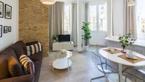 Un saló modern amb un sofà marró, una taula de centre de vidre, una taula de menjador blanca amb cadirees, un televisor i una finestra gran amb cortines.