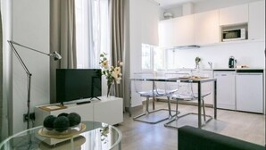 A modern living room with a flat-screen TV, a glass coffee table with decorative items, a dining area with clear chairs, and a kitchen with white cabinets and a microwave.