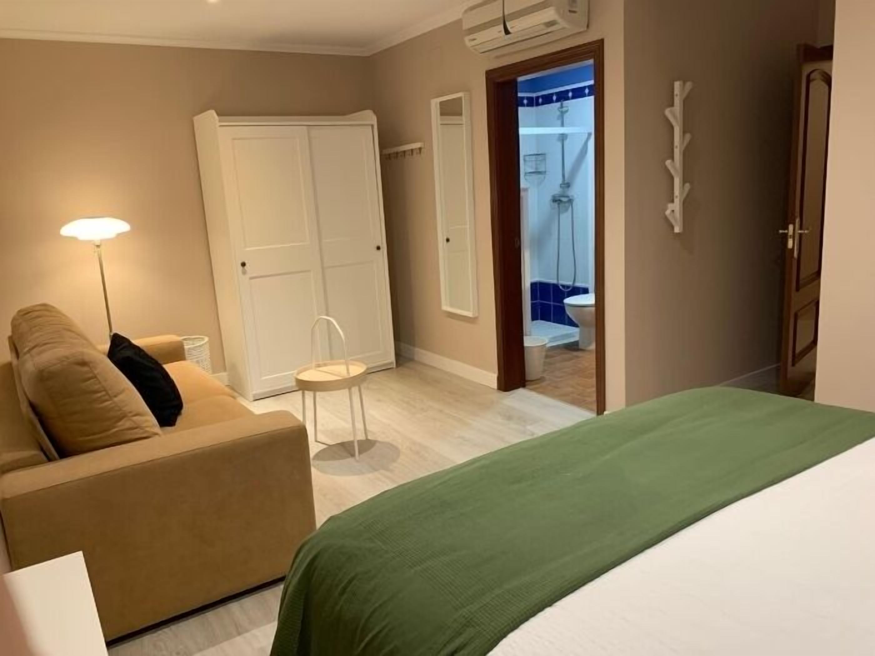 A hotel room with a bed, a sofa, a small table, a mirror, and a door leading to a bathroom.