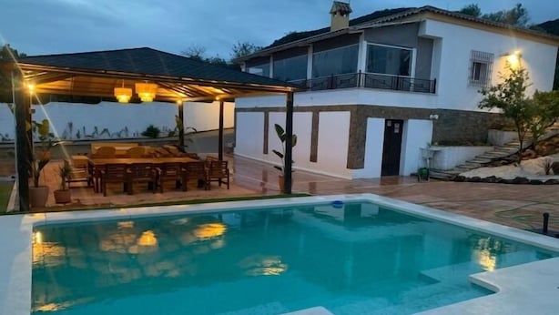 A swimming pool with a covered patio area, a dining table, and a house in the background.