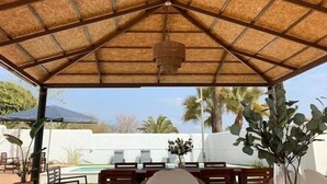 A covered outdoor area with a wooden roof, tables, chairs, and potted plants.