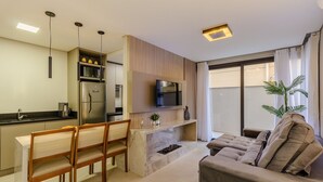 A modern kitchen with a dining area, a flat-screen TV mounted above the fireplace, and a comfortable seating area with a sofa and a potted plant. - DLH Aptos no Coração de Gramado (Gramado)