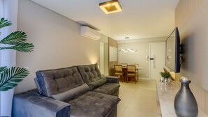 A modern living room with a sofa, a dining area, and a flat-screen TV mounted on the wall. - DLH Aptos no Coração de Gramado  (Gramado)