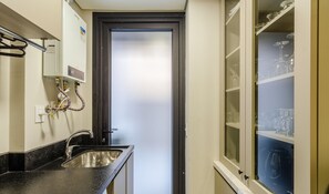 Cocina compacta con encimera negra, pileta de acero inoxidable y puerta de vidrio arenoso que da a una habitación con un armario de vidrio que contiene copas de vino.