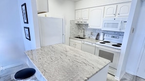 Une cuisine moderne avec des meubles blancs, un plan de travail en granit et un four encastré. On y trouve deux tabourets de bar avec des assises noires.