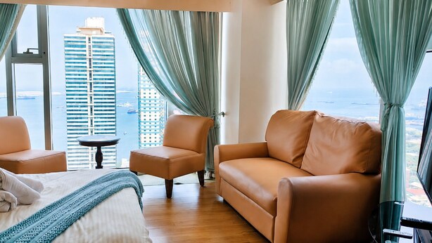 A hotel room with a bed, two armchairs, a sofa, a small table, and a TV. The room has large windows with curtains, a view of a tall building and the sea. - Triple view Condotel Superior manila bay Downtown /st lukes /steps robinson mall (Manila)