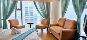 A hotel room with a bed, two armchairs, a sofa, a small table, and a TV. The room has large windows with curtains, a view of a tall building and the sea. - Triple view Condotel Superior manila bay Downtown /st lukes /steps robinson mall (Manila)