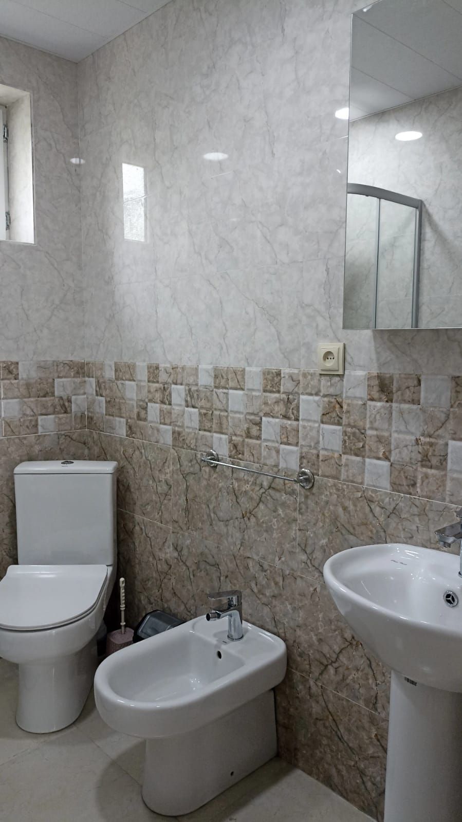 Un baño con inodoro, bidé y lavamanos, paredes de mármol y un espejo.