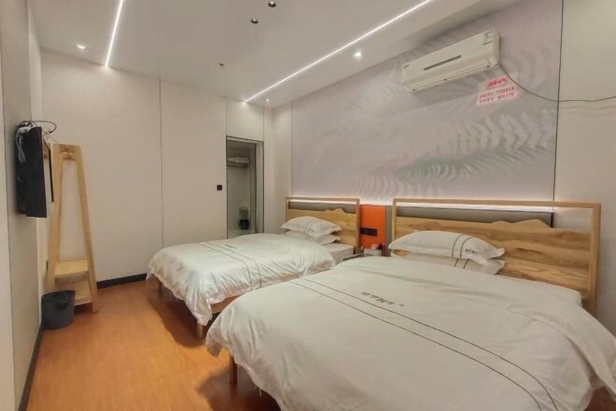 A hotel room with two beds, a wooden floor, a wall-mounted air conditioner, and a small table with a lamp.