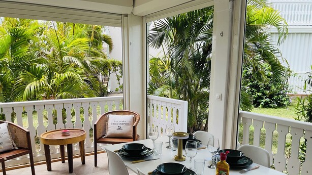 Un espace extérieur abrité, aménagé pour prendre les repas, avec une table dressée pour deux personnes, entourée de chaises, d’une rambarde blanche et d’une végétation luxuriante en arrière-plan.