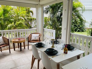Uma área de refeições ao ar livre coberta, com uma mesa posta para dois, rodeada por cadeiras, um corrimão branco e vegetação exuberante ao fundo.