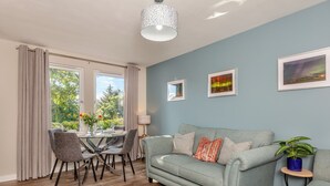 A living room with a grey sofa, dining table, chairs, and a view of greenery through the window. - 2 bedroom accommodation in Nairn (Nairn)