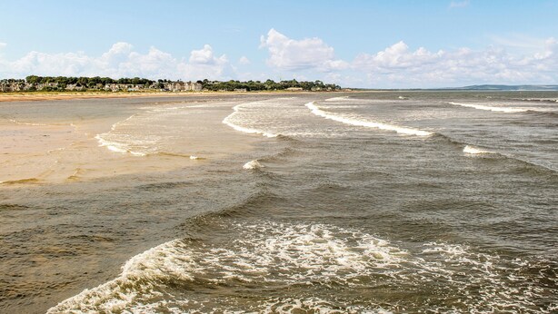 A beach with waves, a clear sky, and a distant shoreline with buildings. - 2 bedroom accommodation in Nairn (Nairn)