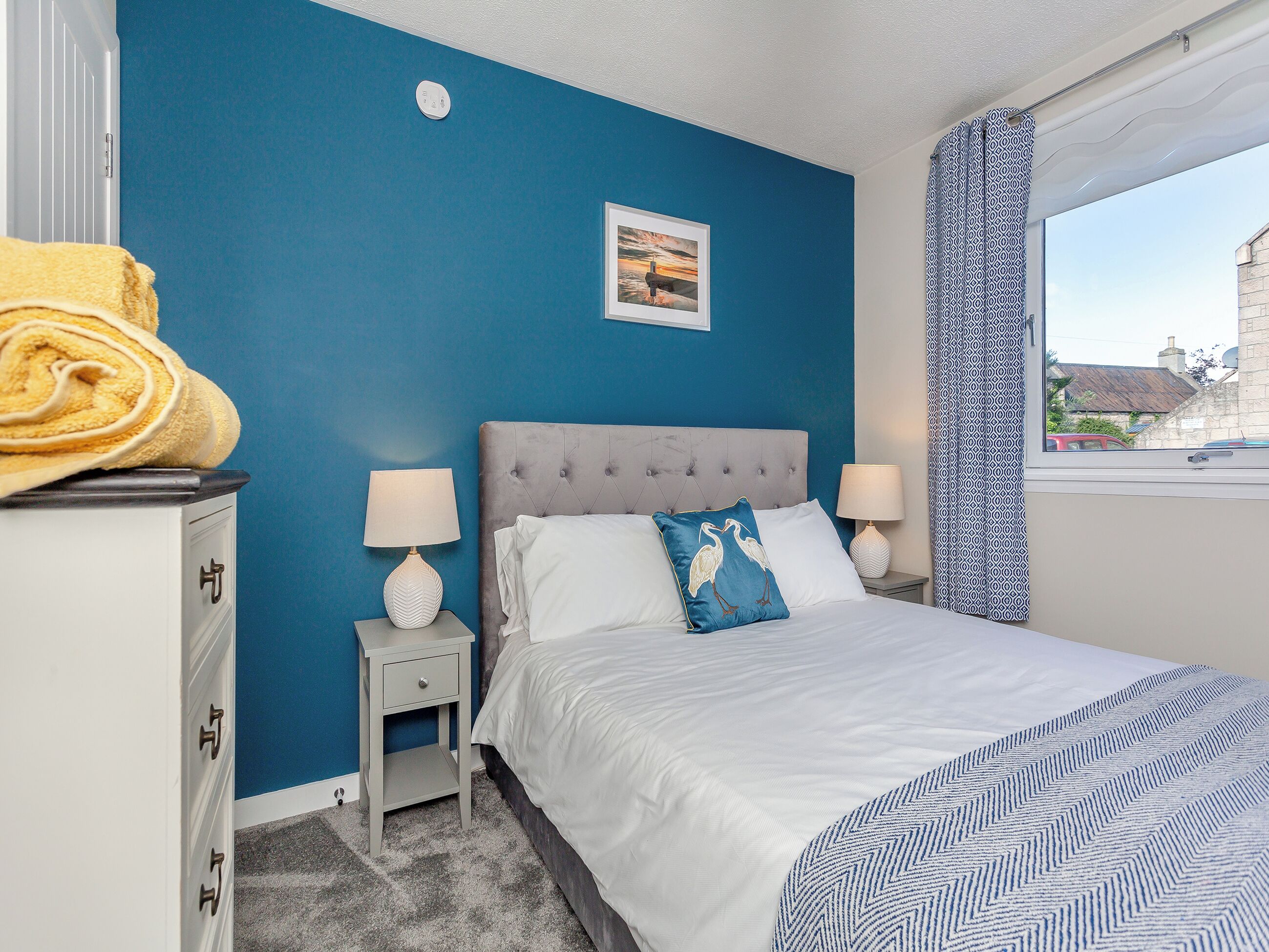 A bedroom with a bed, two bedside tables, a nightstand, a window with curtains, and a wall-mounted picture.