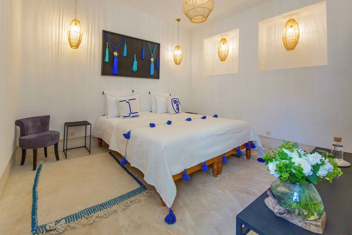 A bedroom with a bed, a chair, a small table with a vase of flowers, and wall-mounted lights.