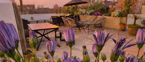 A rooftop patio with a table and chairs, surrounded by potted plants and flowers.