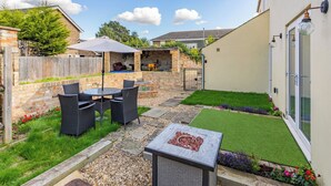 Une terrasse avec une table et des chaises, un espace barbecue et un petit jardin agrémenté de gazon synthétique et de fleurs.