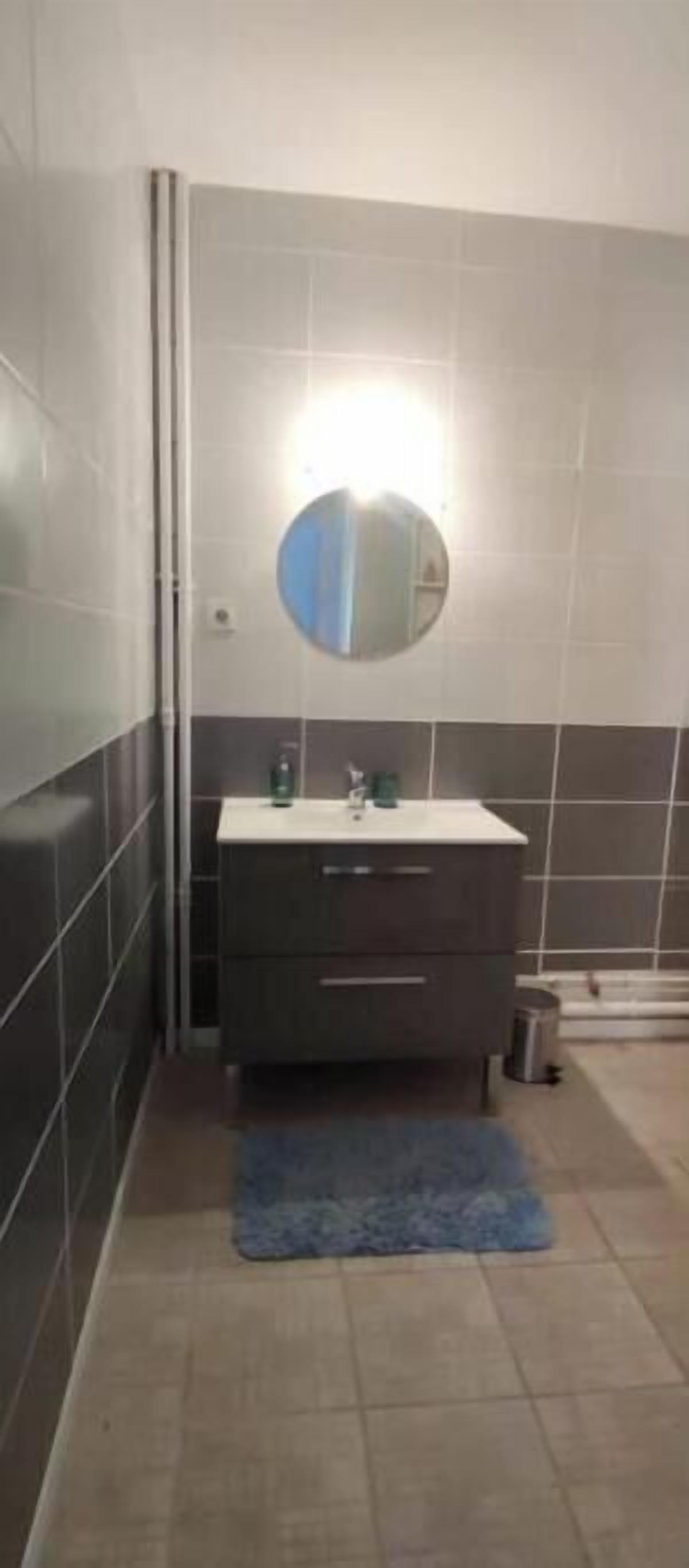 Un bagno con un lavandino bianco, piastrelle scure, uno specchio rotondo e un tappeto blu.