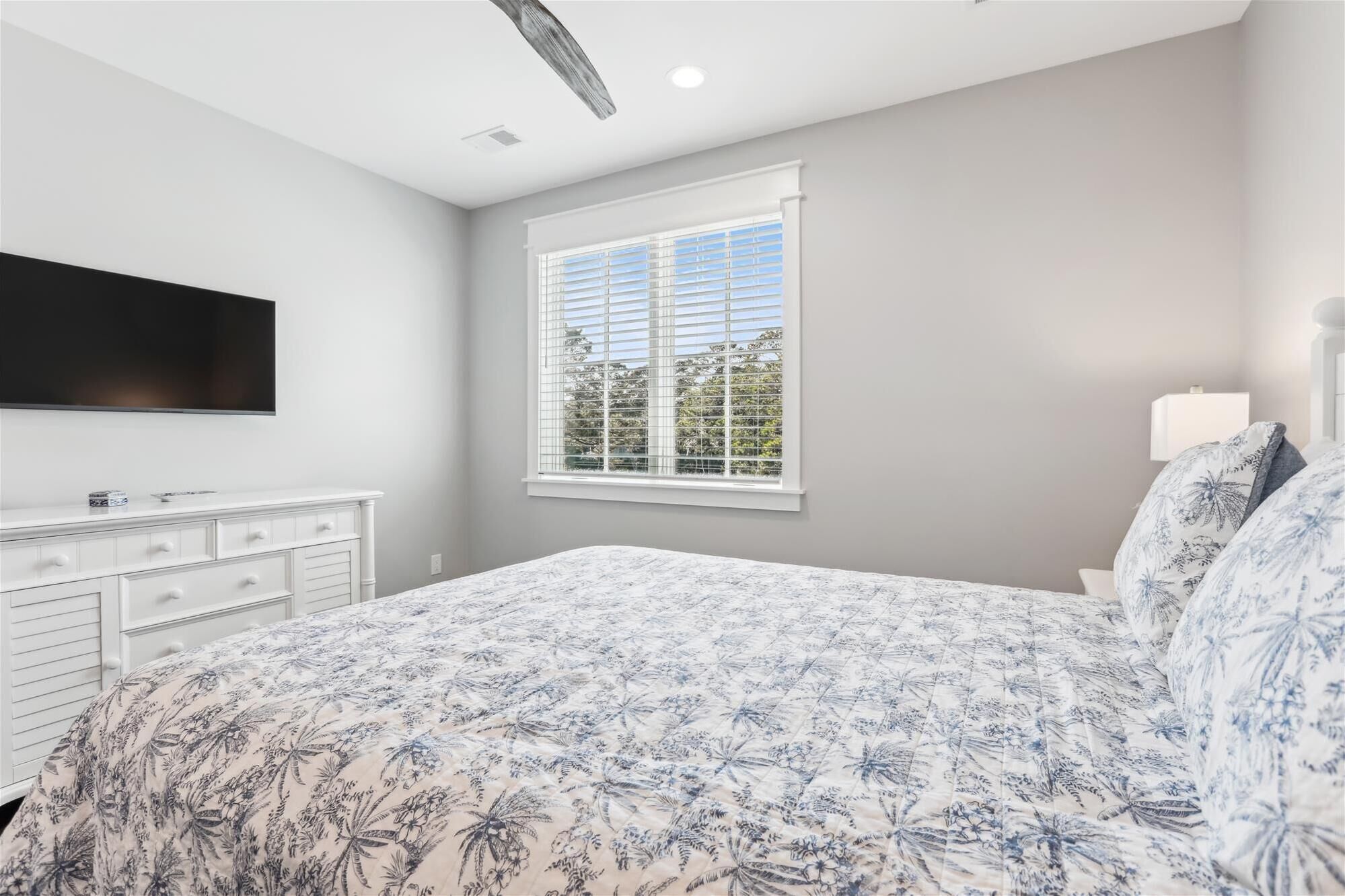 A bedroom with a bed, a dresser, a TV mounted on the wall, and a window with blinds.