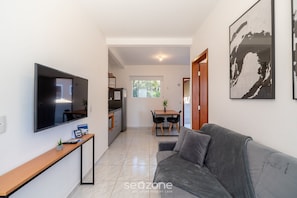 A modern living room with a flat-screen TV, a grey sofa, a dining area with a table and chairs, and a kitchen in the background. - JMA Apartamentos em Jurere (Florianópolis)