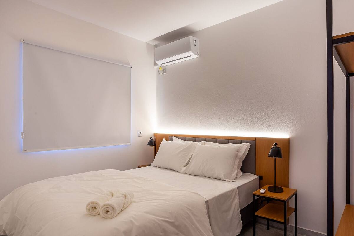 A hotel room with a bed, a nightstand, a lamp, a hanging clothes rack, and an air conditioning unit.