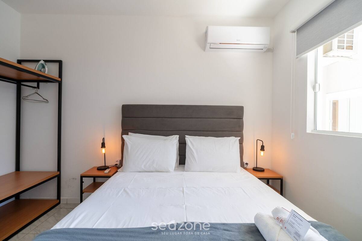 A neatly arranged hotel room with a bed, bedside tables, a hanging shelf, and an air conditioning unit.