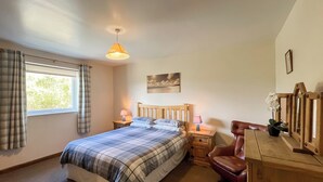A bedroom with a bed, two bedside tables with lamps, a wooden chair, a dresser, a window with curtains, and a painting on the wall.