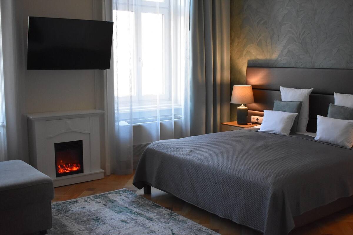Een slaapkamer met een bed, een aan de muur bevestigde televisie, een open haard en een nachtkastje met een lamp.