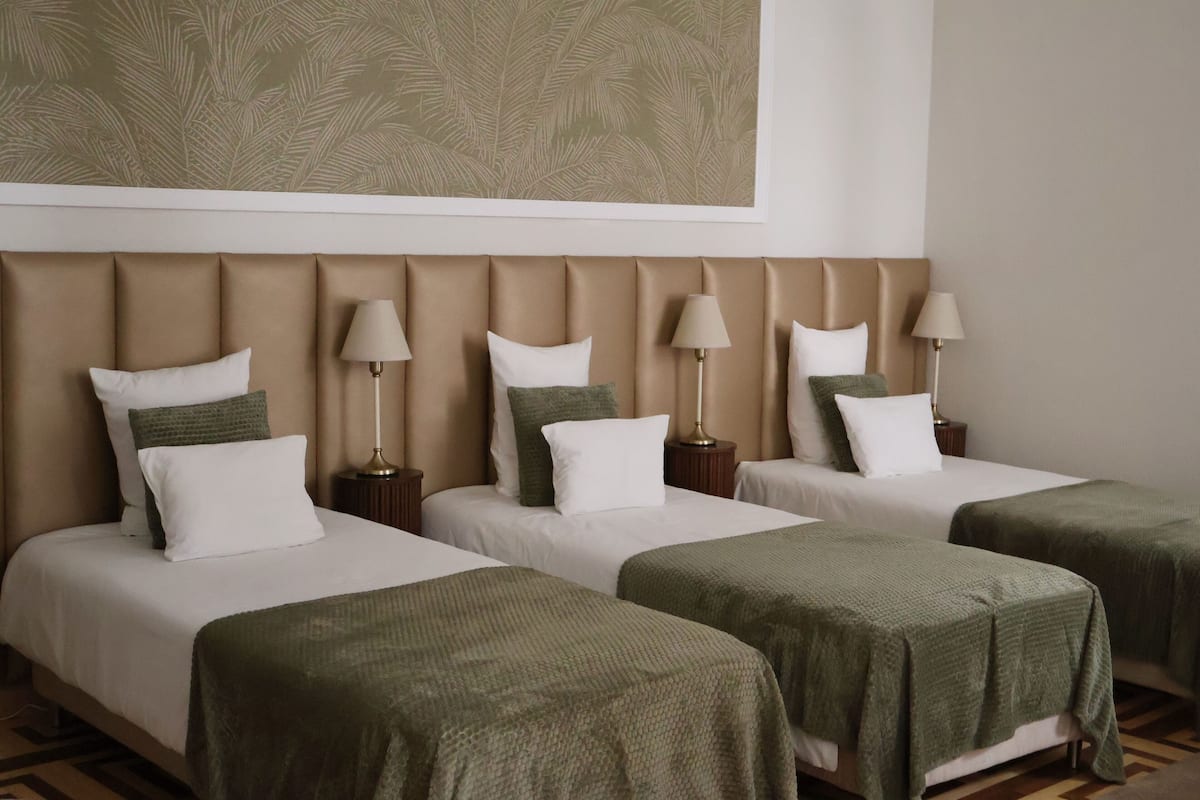 Een hotelkamer met drie bedden, elk met een groene deken en witte kussens. Er staan twee bedlampjes op houten nachtkastjes.