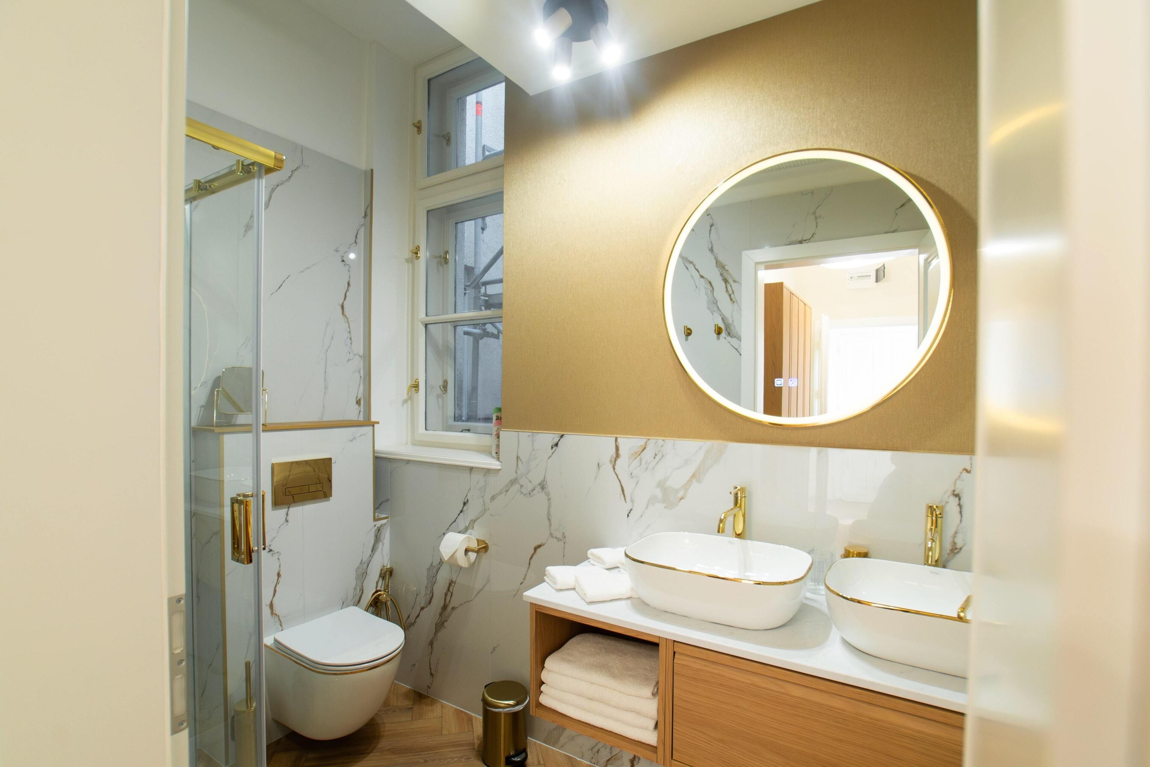 Une salle de bain moderne avec une douche à parois de verre, des toilettes blanches et deux lavabos dotés de robinetterie dorée.