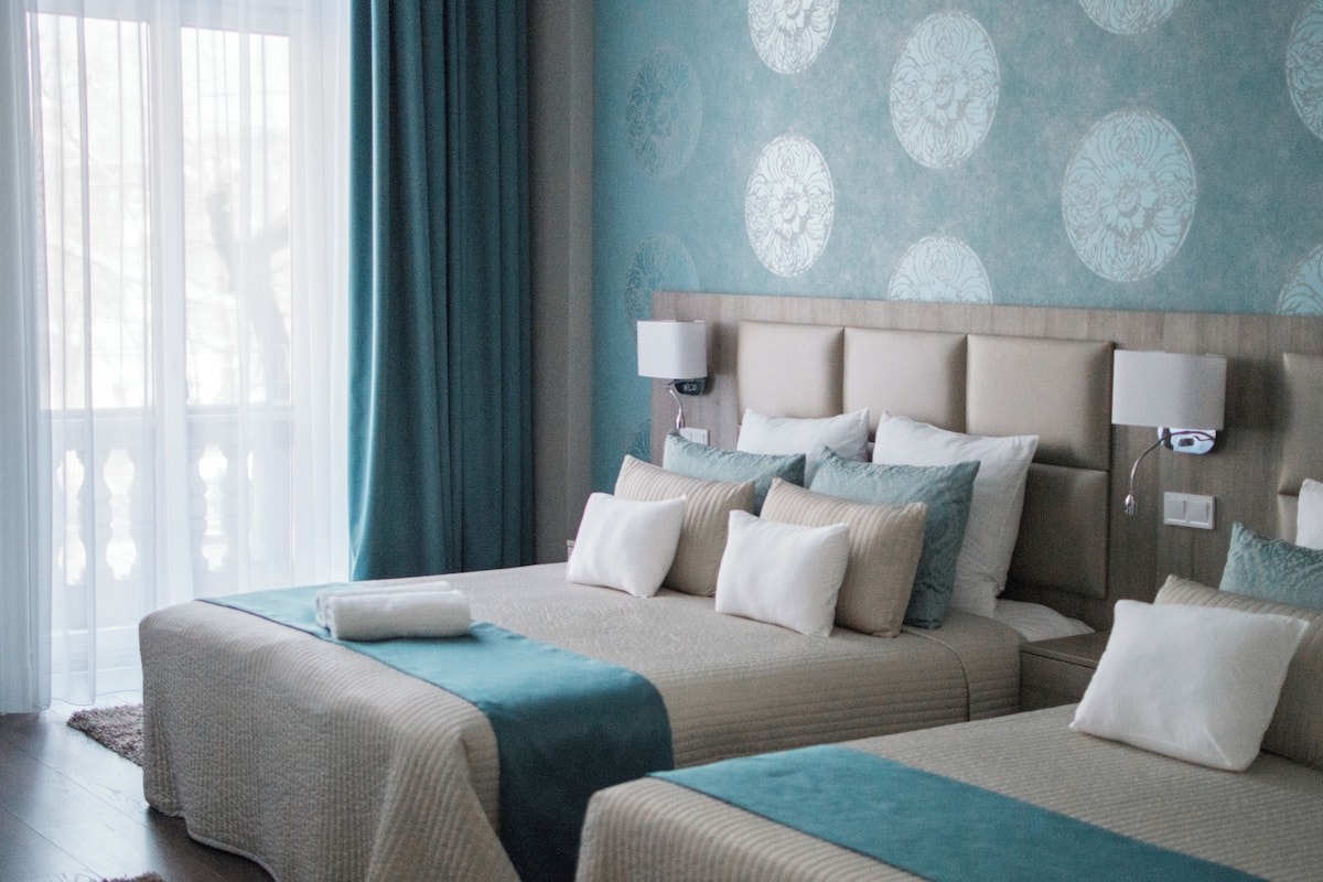 Een hotelkamer met twee bedden, een groot raam met gordijnen, een behang met patroon en bedlampjes.