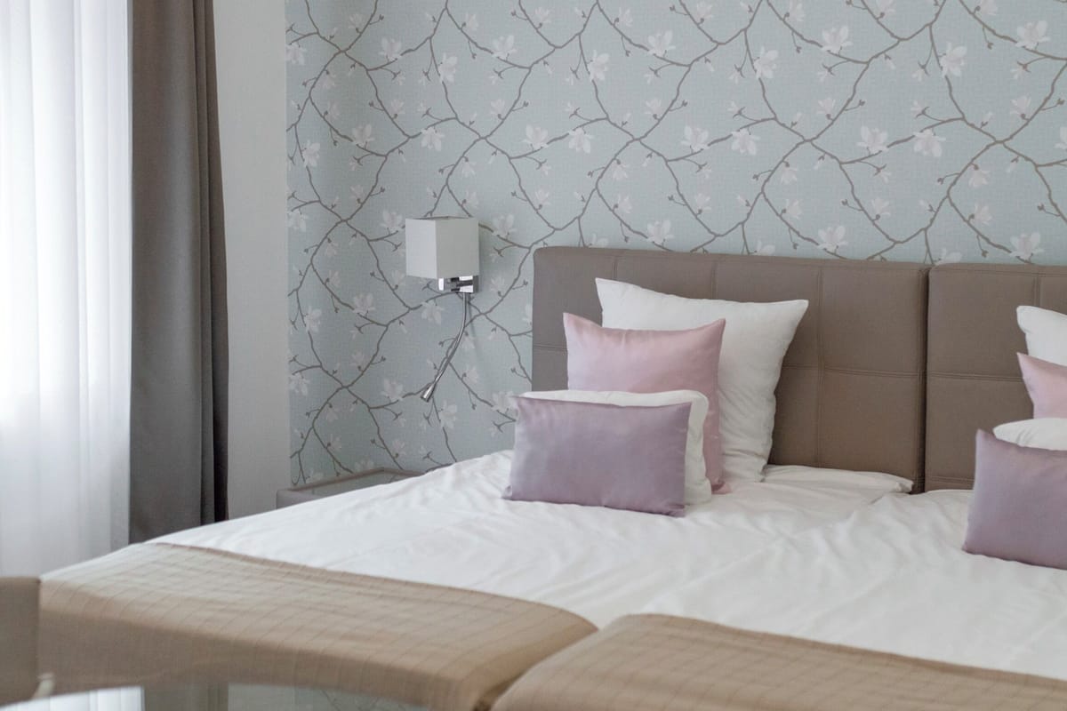Een hotelkamer met een bed, twee kussens, een nachtkastje en een wand met bloemenprint.
