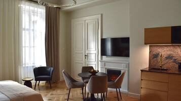 A hotel room with a bed, a dining table with chairs, a fireplace, a TV mounted on the wall, and a large window with curtains.