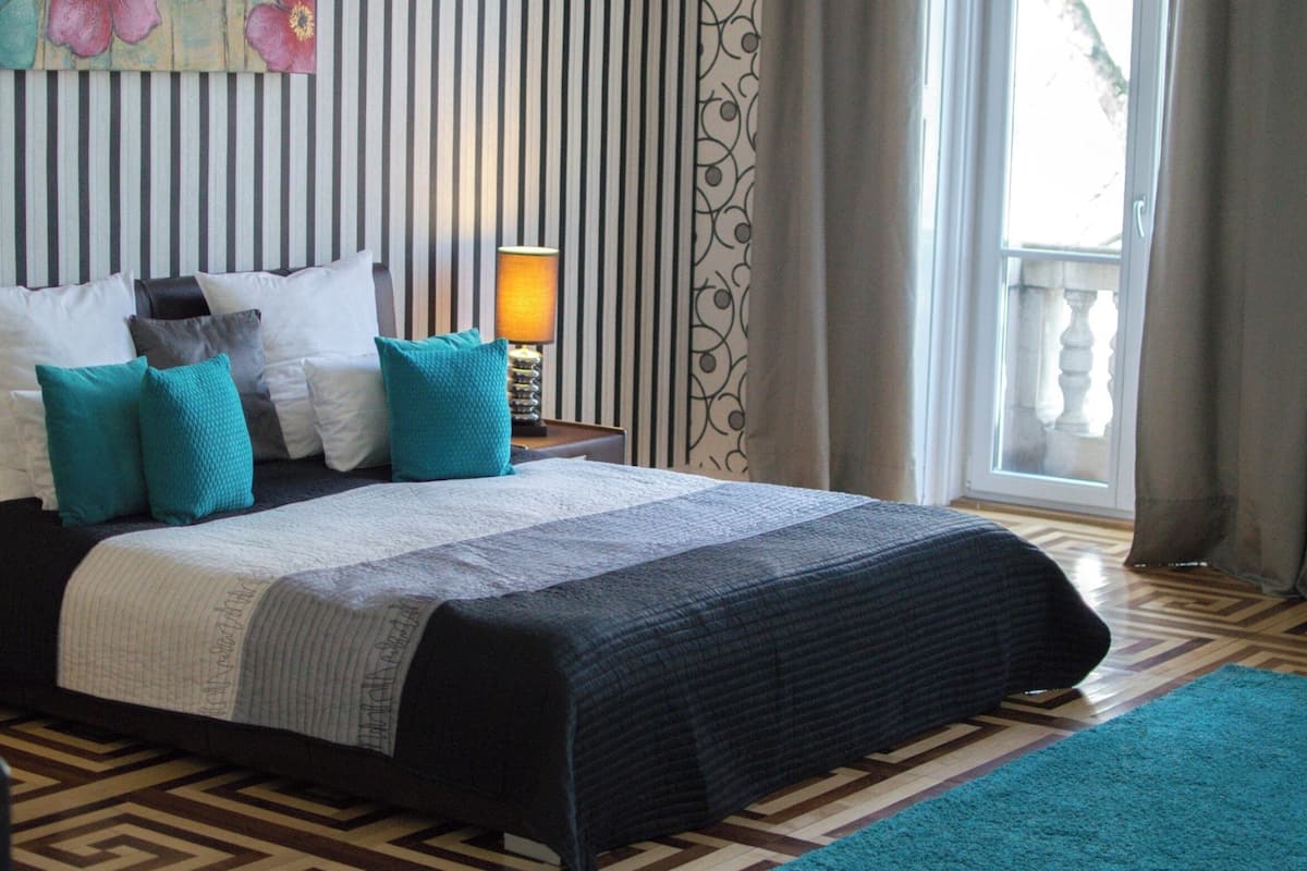 Een net opgemaakt bed met een gestreept hoofdbord, een bedlampje en uitzicht op de omgeving door een raam met gordijnen.