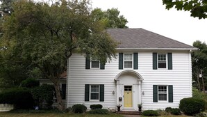 A two-story white house with green shutters, a yellow front door, and a small front yard with a pathway leading to the entrance.