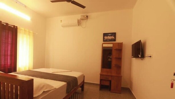 A small, simple room with a bed, a wooden desk, a chair, a television, and a mirror. - Periyar Homes Deluxe Room 102 (Kumily)