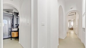 Un couloir moderne aux murs blancs et au sol de couleur claire, qui mène à une salle de bain équipée d’un lavabo et d’un miroir.
