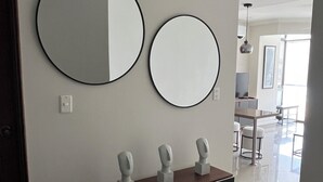 Un couloir moderne avec deux miroirs ronds accrochés au mur, une console en bois foncé ornée de trois bustes sculptés, et un sol carrelé et lumineux qui mène à un espace repas.