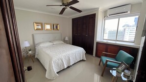 Une chambre d’hôtel avec un lit, un ventilateur de plafond, une fenêtre donnant sur la ville et une petite table avec une plante.