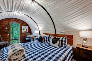 Een gezellig ingerichte tent met een geruit bed, een houten stoel, een tafeltje en een lamp.