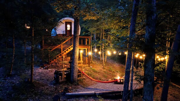 A rustic cabin with a wooden staircase leading to a lofted bedroom, surrounded by trees and string lights.