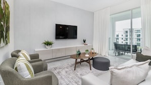 A modern living room with a flat-screen TV, grey sofas, a coffee table, and a balcony with a view of buildings.