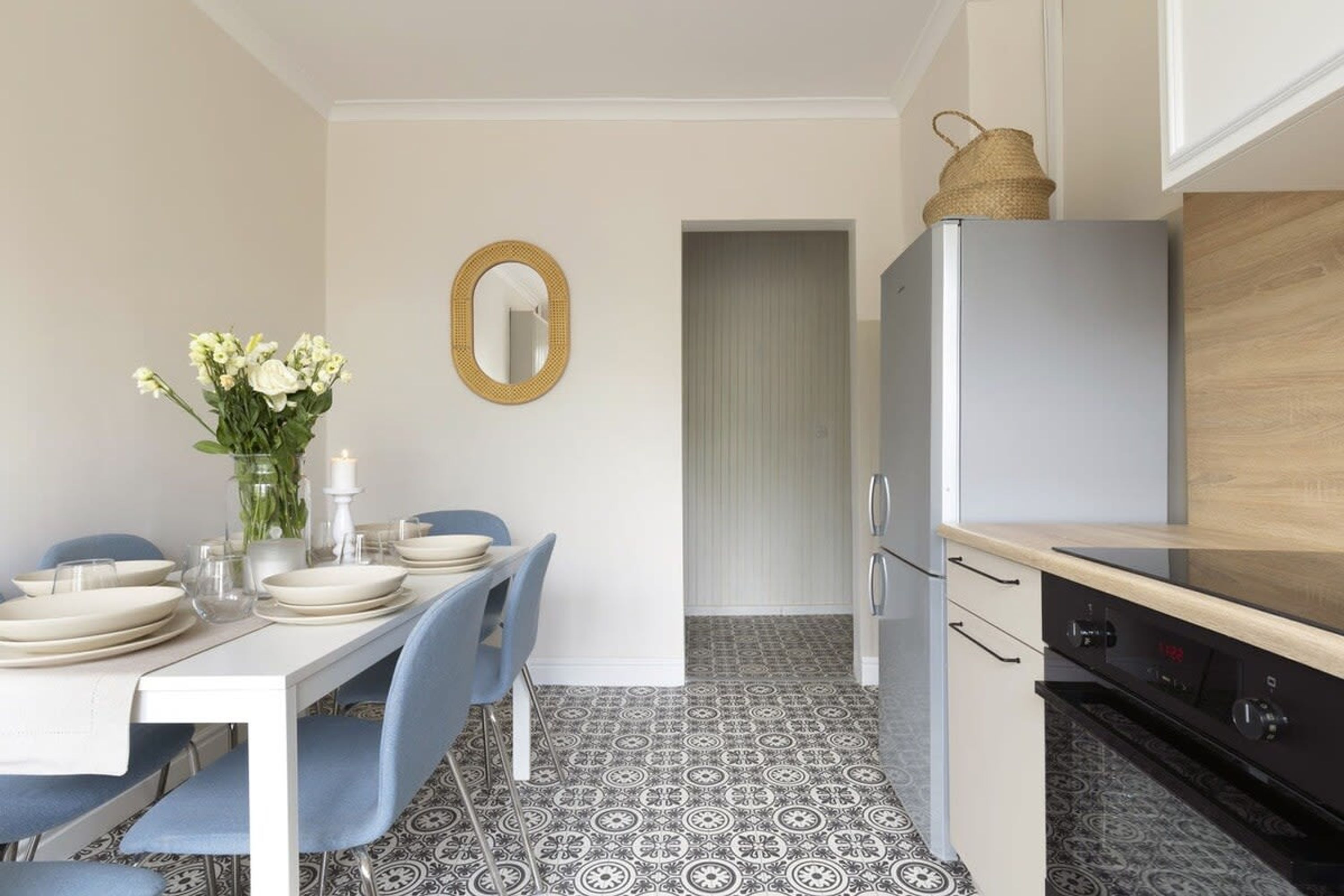 A compact kitchen with a modern refrigerator, a dining table set with plates and a vase of flowers, and patterned floor tiles.