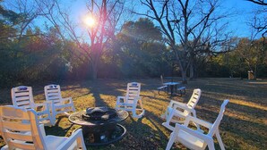 A patio with white plastic chairs arranged around a fire pit, surrounded by trees and a clear sky. - Cozy Camper 2-bedroom RV in Americus GA  (Americus)