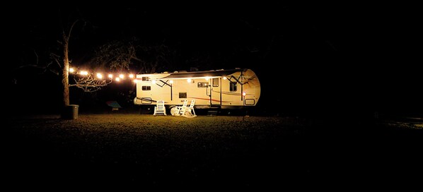 A well-lit RV with a ladder and a small table in front, surrounded by trees. - Cozy Camper 2-bedroom RV in Americus GA (Americus)